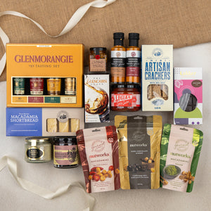 The Hamper Boutique Co’s Glenmorangie Ultimate Tasting Hamper features gourmet whisky, sauces, macadamia nuts, crackers, shortbread, chocolate, honey, and preserves—all elegantly arranged with ribbon and fabric accents on a beige surface.