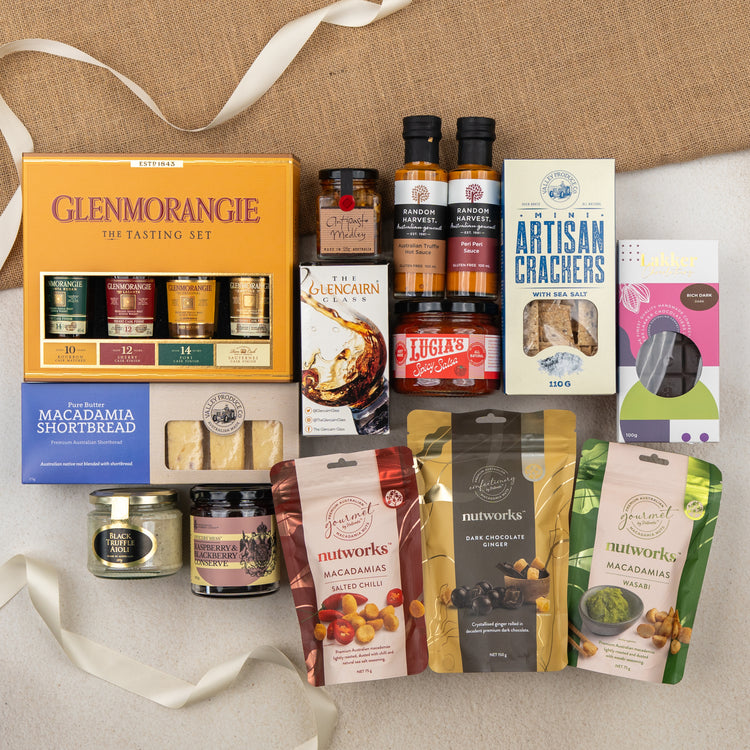 The Hamper Boutique Co’s Glenmorangie Ultimate Tasting Hamper features gourmet whisky, sauces, macadamia nuts, crackers, shortbread, chocolate, honey, and preserves—all elegantly arranged with ribbon and fabric accents on a beige surface.
