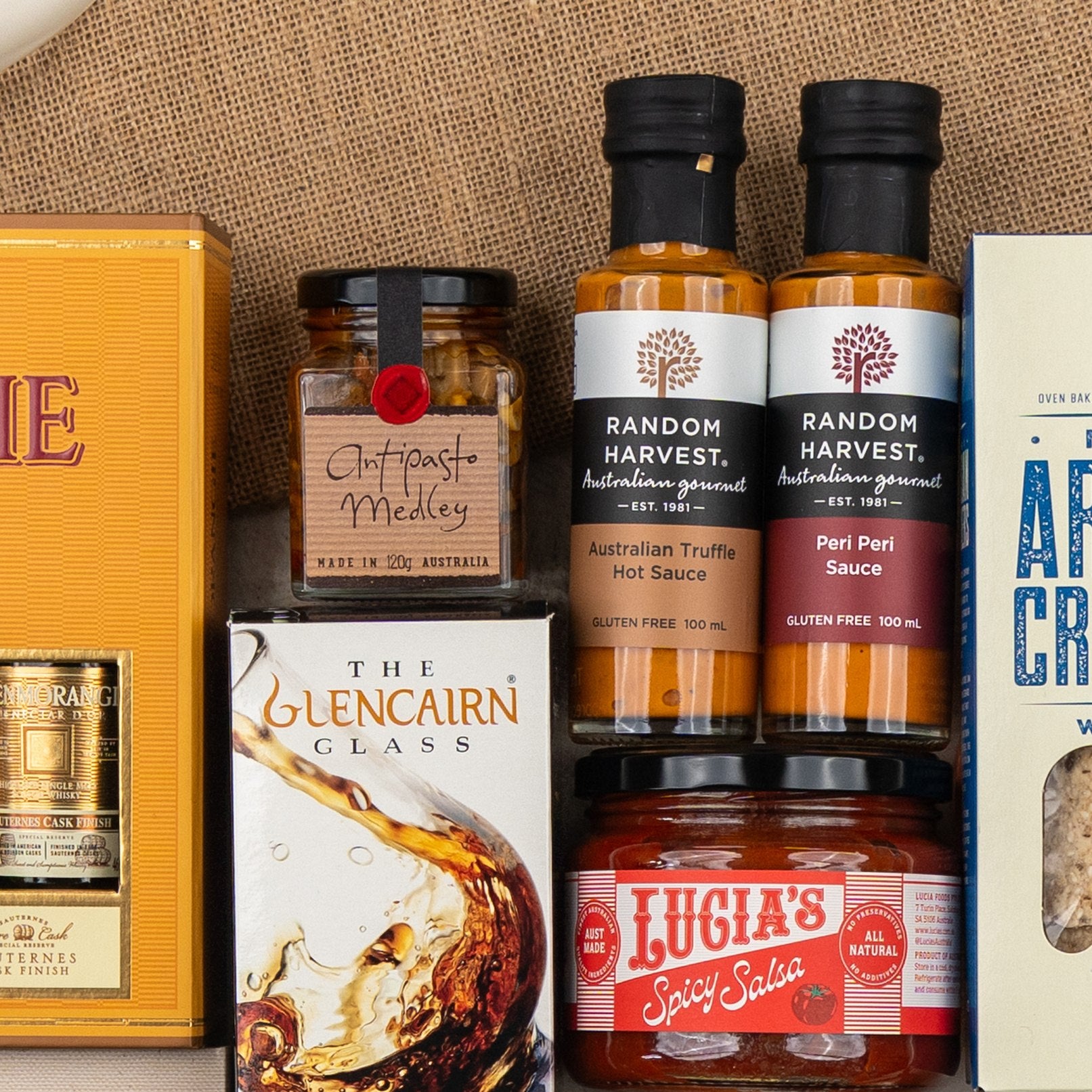 The Hamper Boutique Co's Glenmorangie Ultimate Tasting Hamper features gourmet sauces, antipasto medley, spicy salsa, crackers, and a Glencairn whisky glass, all elegantly arranged on a burlap surface.
