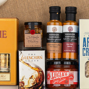 The Hamper Boutique Co's Glenmorangie Ultimate Tasting Hamper features gourmet sauces, antipasto medley, spicy salsa, crackers, and a Glencairn whisky glass, all elegantly arranged on a burlap surface.