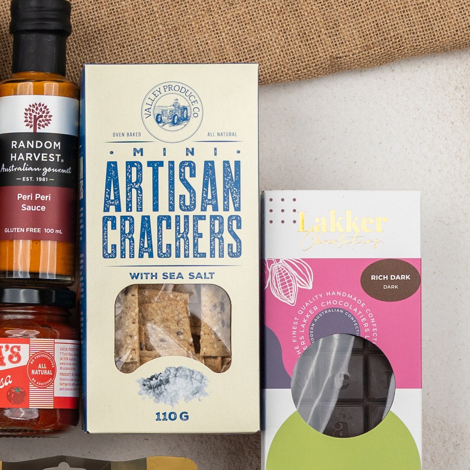 A close-up of The Hamper Boutique Co's Glenmorangie Ultimate Tasting Hamper, featuring peri peri sauce, sea salt mini artisan crackers, and rich dark chocolate, arranged on a light surface with burlap in the background.