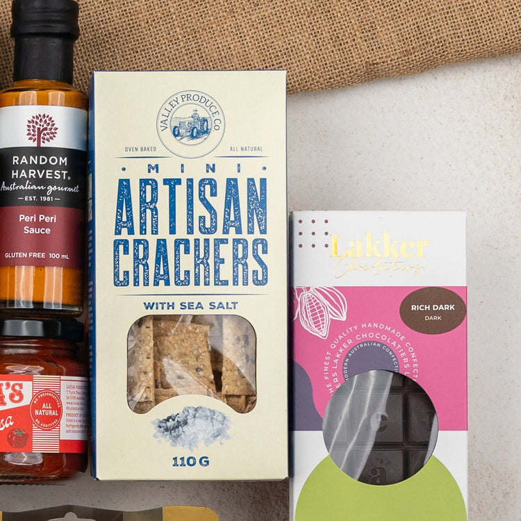 A close-up of The Hamper Boutique Co's Glenmorangie Ultimate Tasting Hamper, featuring peri peri sauce, sea salt mini artisan crackers, and rich dark chocolate, arranged on a light surface with burlap in the background.