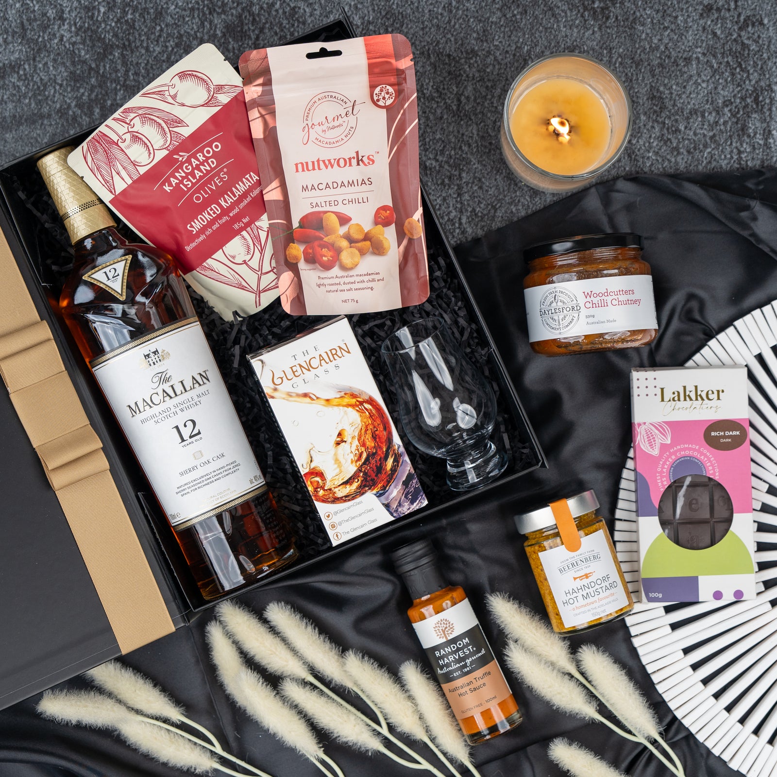 The Macallan Indulgence Hamper by The Hamper Boutique Co features The Macallan whisky, Glencairn glasses, salted macadamias, Lakker chocolate, Woodbourne chutney, a soy candle, honey mustard, and a folding fan. Displayed on gray with decorative grasses.