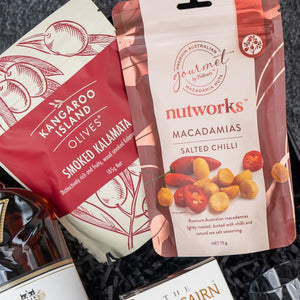 A close-up of the Macallan Indulgence Hamper by The Hamper Boutique Co, featuring packaged snacks like smoked kalamata olives in a red pouch and salted chilli macadamias, displayed elegantly in a gift basket.
