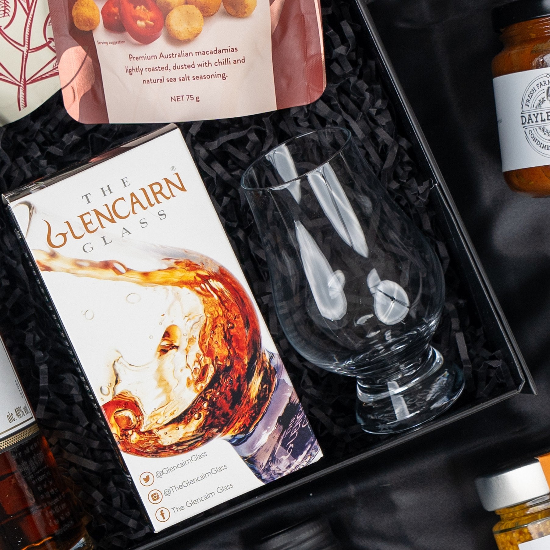 The Hamper Boutique Co's Macallan Indulgence Hamper features a Glencairn whiskey glass in its box, a glass tumbler, macadamia nuts, and preserves, all elegantly arranged on black shredded paper—an ideal gift for any occasion.