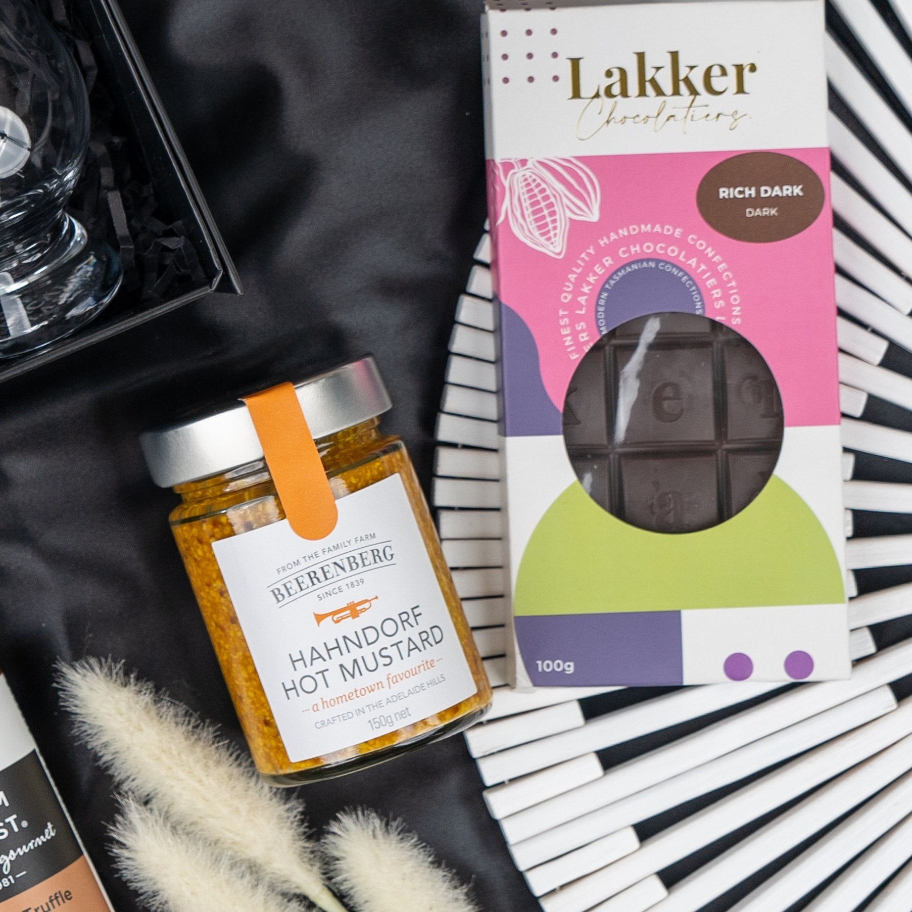 A Macallan Indulgence Hamper by The Hamper Boutique Co is displayed on a black and white striped surface with decorative grass and part of a black gift box visible.