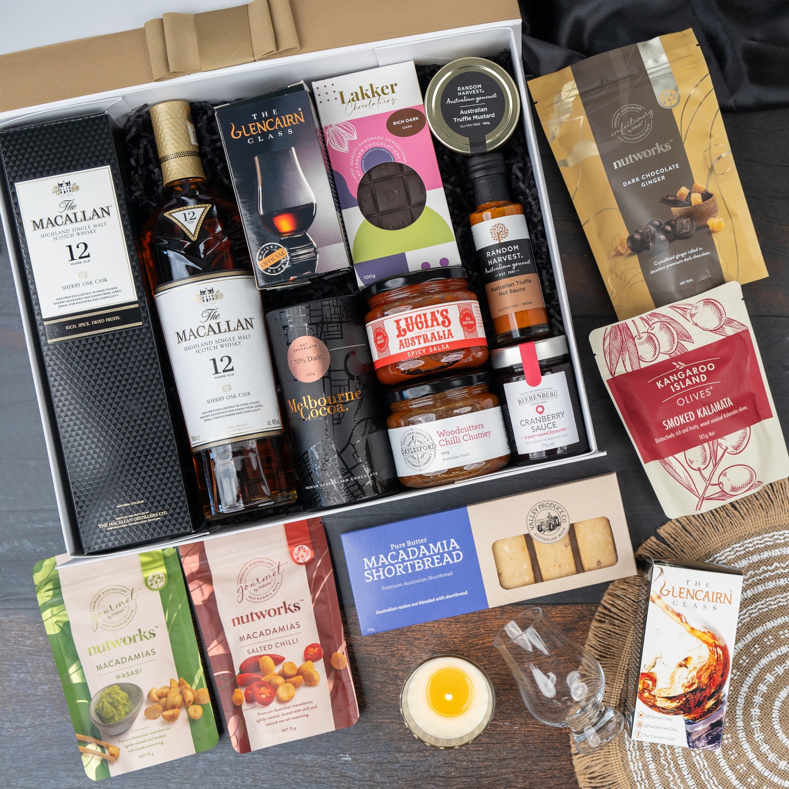 The Macallan Collector's Edition Hamper by The Hamper Boutique Co includes a bottle of The Macallan 12 whisky, Glencairn glass, chocolates, cookies, savory snacks, nuts, a candle, and gourmet preserves arranged with extra snacks beside the box.