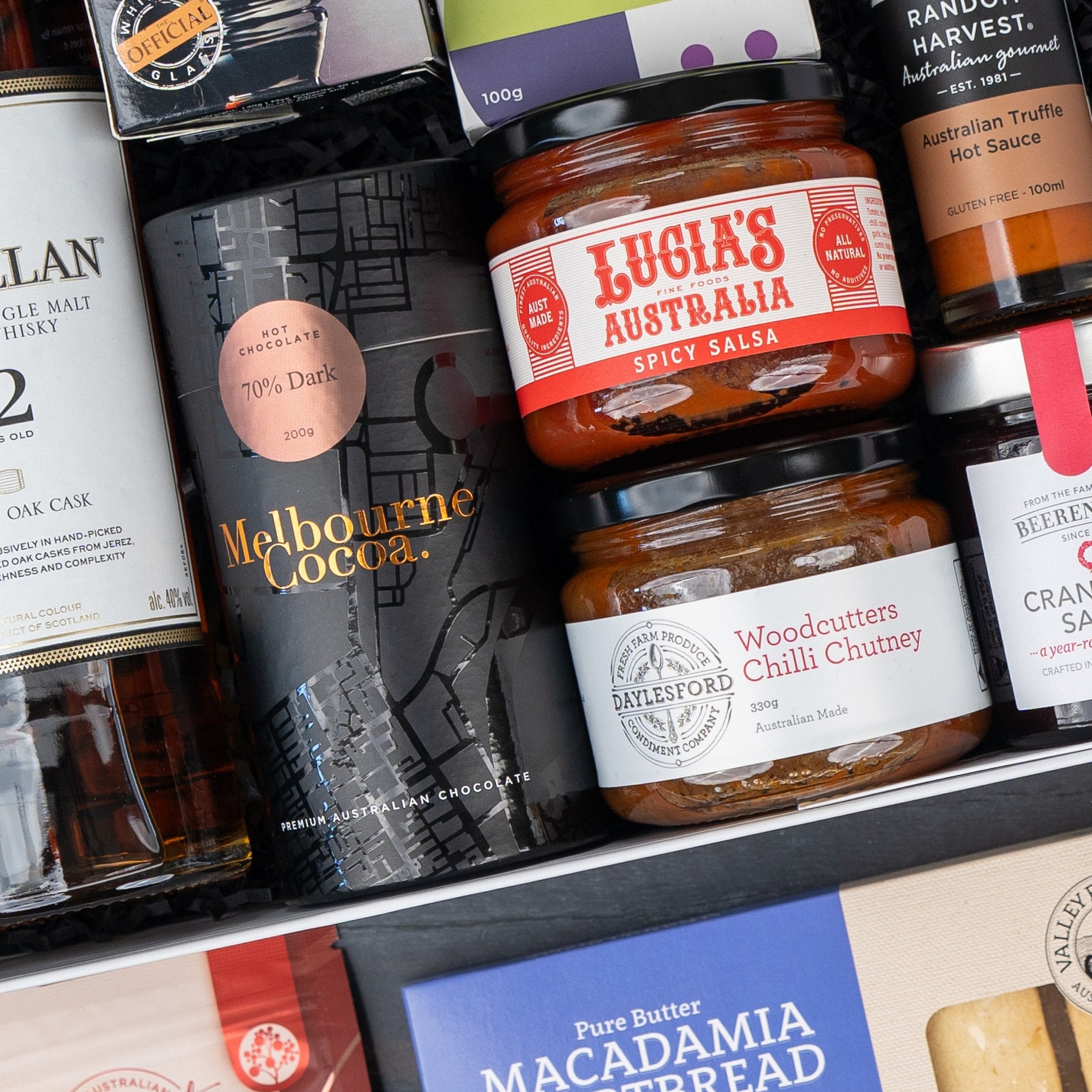 A close-up of The Hamper Boutique Co’s Macallan Collector's Edition Hamper, featuring whisky, Melbourne Cocoa chocolate, Lucia’s spicy salsa, Woodcutters chili chutney, macadamia spread, truffle hot sauce, and cranberry sauce.