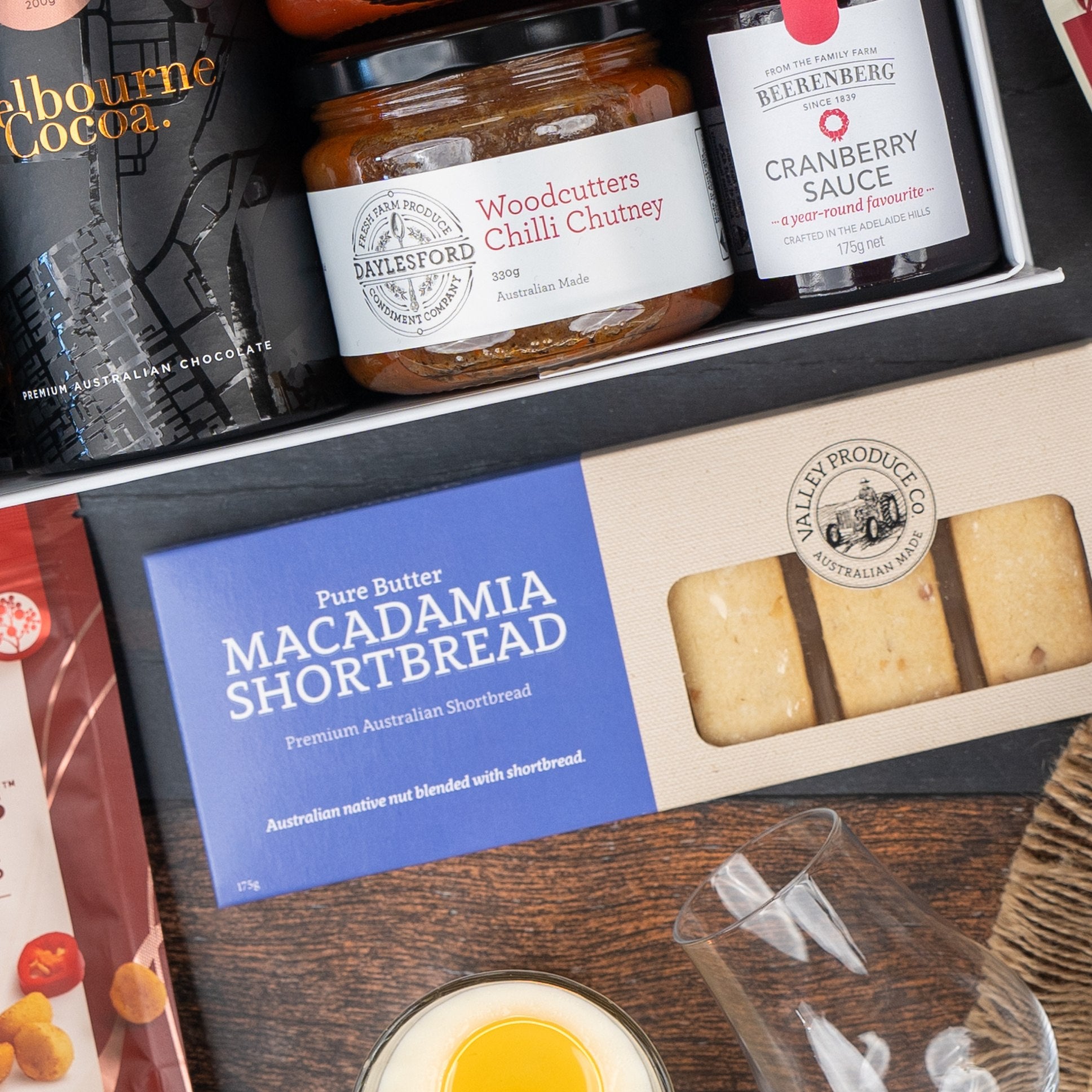 The Hamper Boutique Co's Macallan Collector's Edition Hamper includes macadamia shortbread, sparkling red grape juice, woodcutters chili chutney, cranberry sauce, and a glass, all stylishly arranged on a wooden surface.