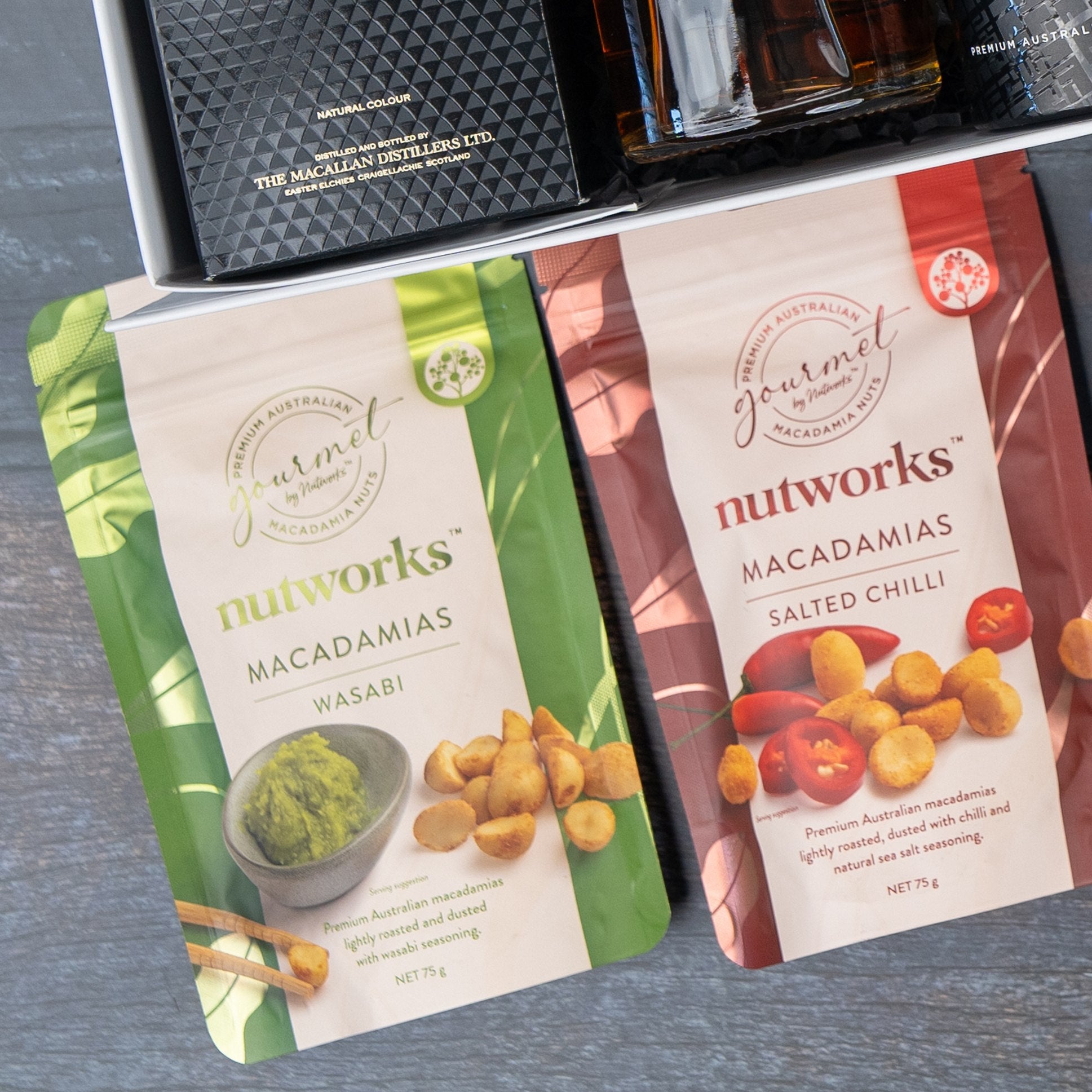 Two packets of Nutworks macadamias (wasabi in green, salted chili in red) are shown beside a Macallan Collector's Edition Hamper by The Hamper Boutique Co, all displayed on a gray surface.