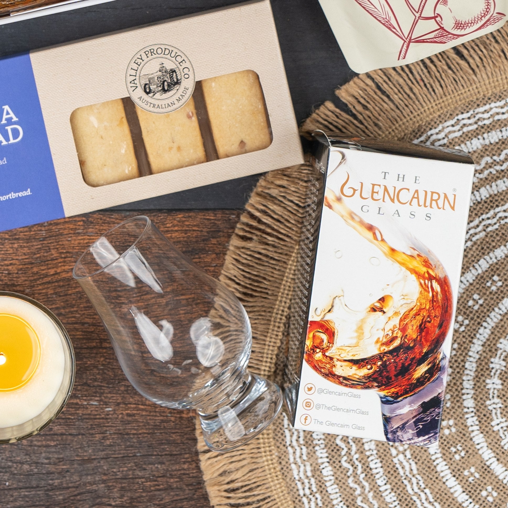 The Macallan Collector's Edition Hamper by The Hamper Boutique Co includes a Glencairn whisky glass in a whisky splash box, Australian shortbread, and a candle, all arranged on a woven placemat atop a wooden surface.