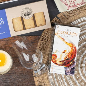 The Macallan Collector's Edition Hamper by The Hamper Boutique Co includes a Glencairn whisky glass in a whisky splash box, Australian shortbread, and a candle, all arranged on a woven placemat atop a wooden surface.