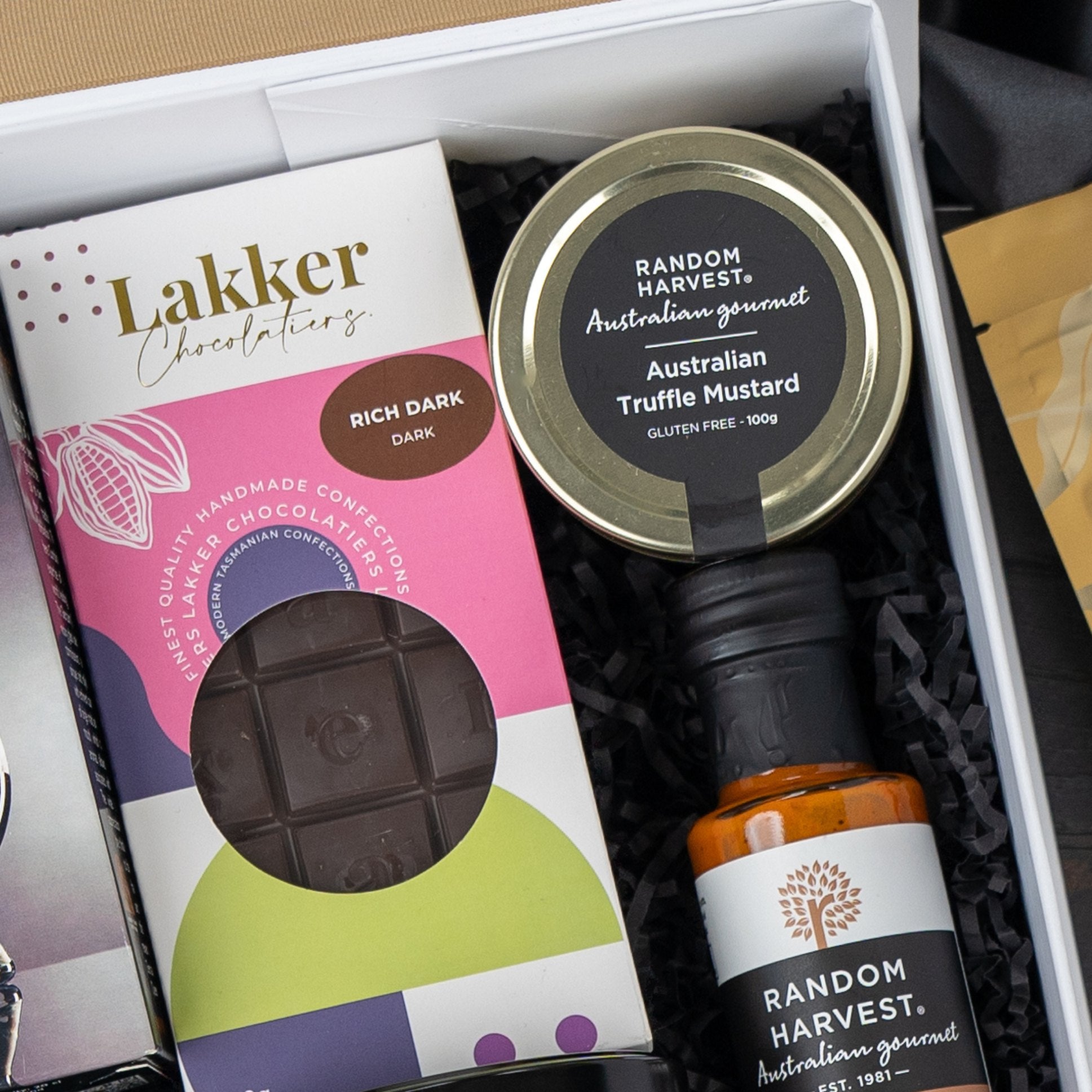The Macallan Collector's Edition Hamper by The Hamper Boutique Co includes a Lakker Chocolatiers rich dark chocolate bar, Random Harvest Australian Truffle Mustard, and a Random Harvest gourmet condiment, all arranged on black paper shred.