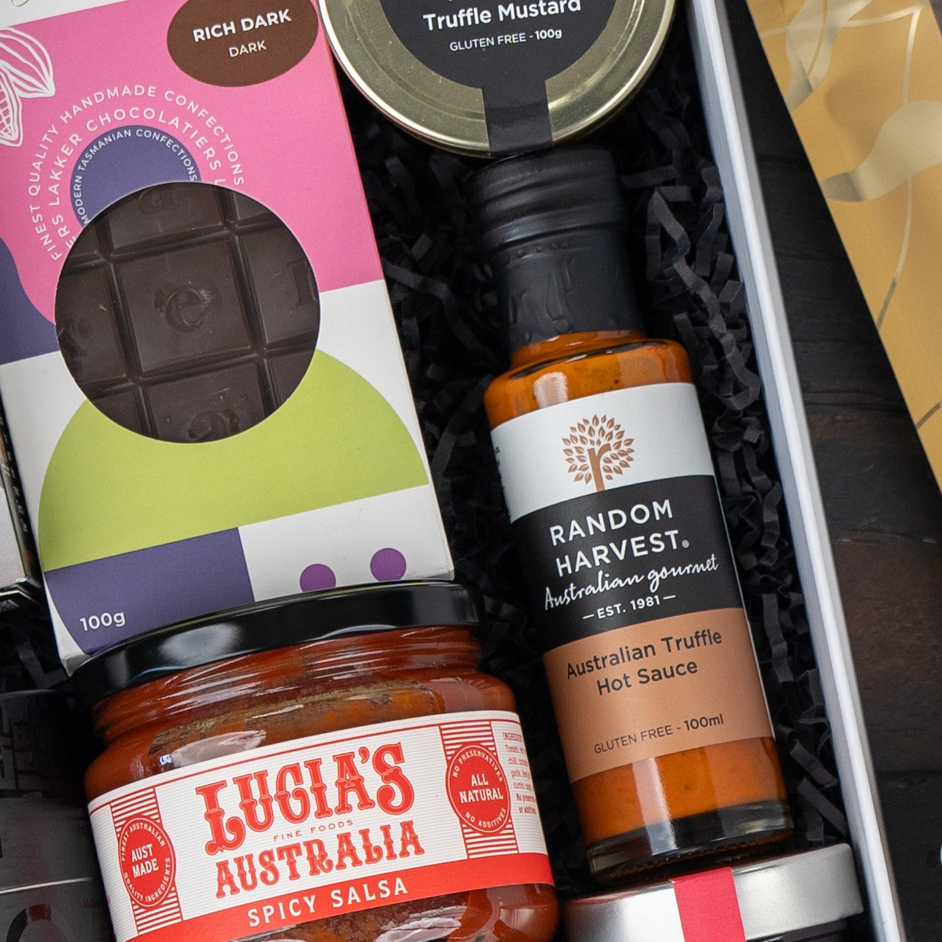 A close-up of The Hamper Boutique Co’s Macallan Collector's Edition Hamper featuring gourmet treats: a chocolate bar, truffle mustard jar, Australian truffle hot sauce bottle, and Lucia’s Australia spicy salsa jar.