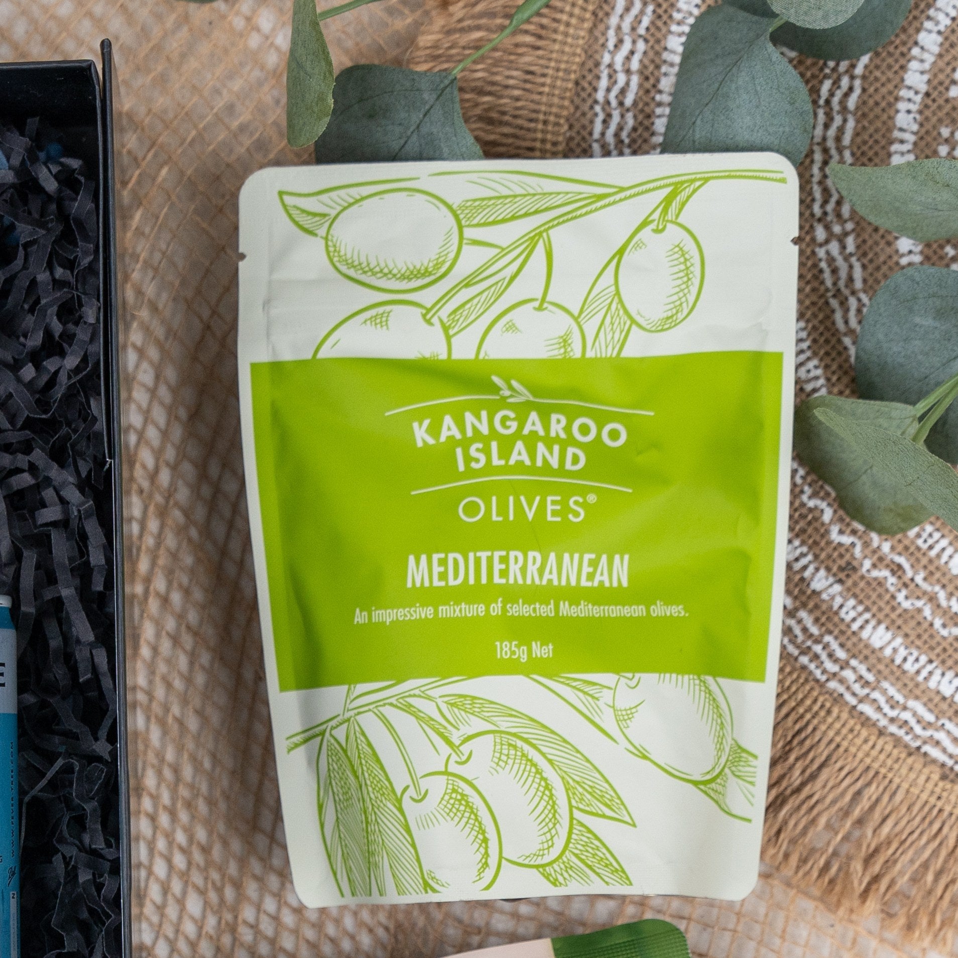 A green and white pouch labeled Tanqueray Delights Hamper by The Hamper Boutique Co sits on a woven surface next to greenery and a black box. The pouch features olive illustrations and contains 185g of Mediterranean olives.