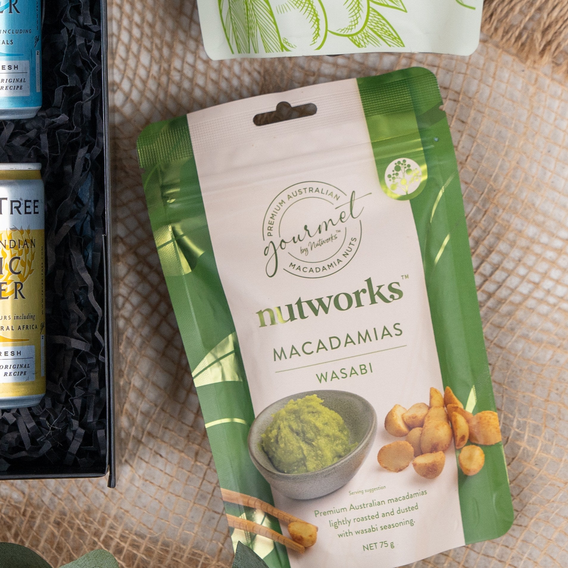 A Tanqueray Delights Hamper from The Hamper Boutique Co is displayed on a woven mat, next to other partially visible food packaging. The hamper's package is prominently shown in the arrangement.