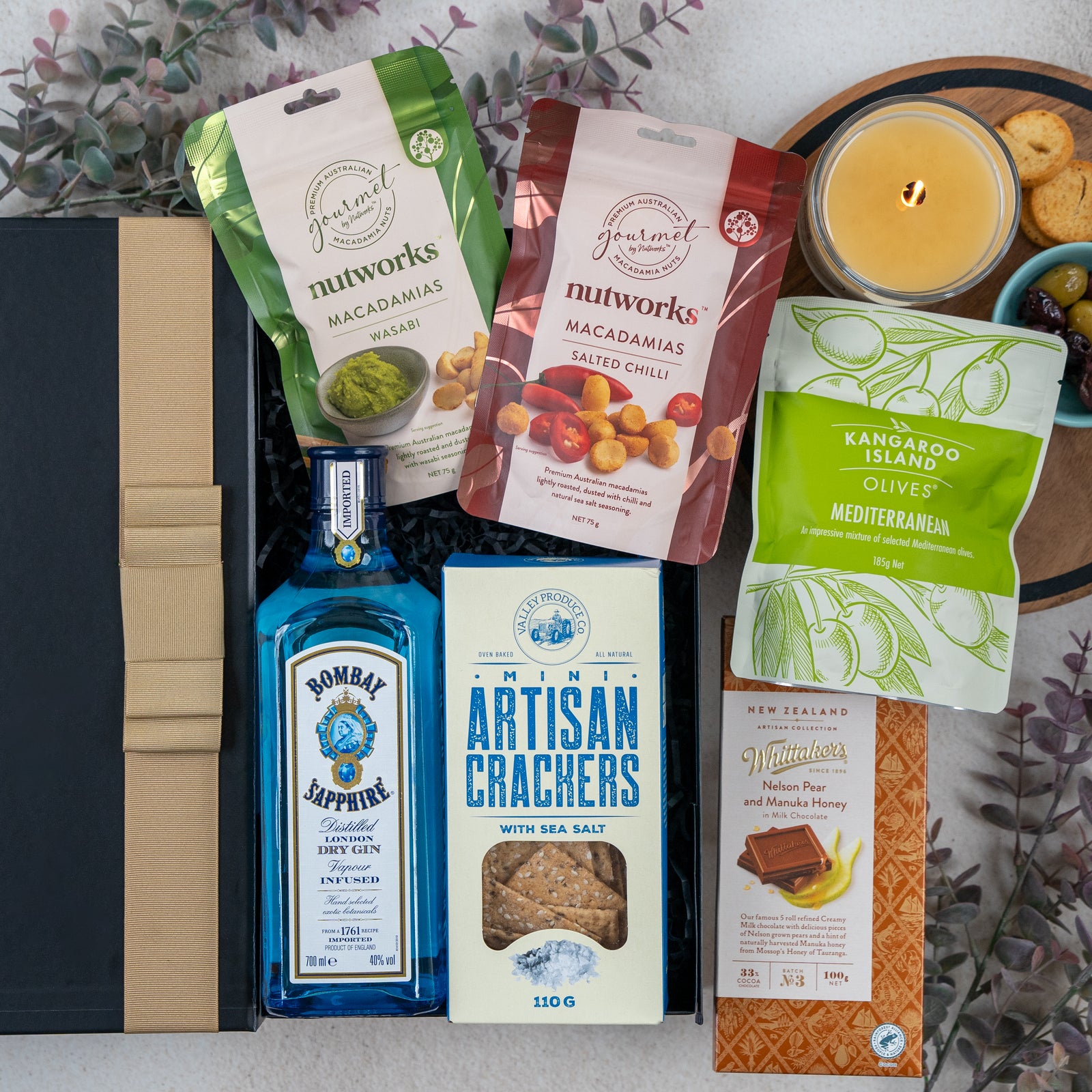 The Hamper Boutique Co's Bombay Sapphire Botanical Escape Hamper features gin, artisan crackers, salt & honey macadamias, Mediterranean olives, a honey chocolate bar, and a yellow candle on a floral-accented decorative surface.