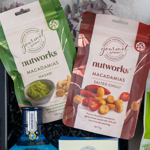 Two Nutworks macadamia nut packets—Wasabi (green) and Salted Chilli (red)—with images of seasoned nuts are displayed above a blue Bombay Sapphire bottle in The Hamper Boutique Co's Botanical Escape Hamper.