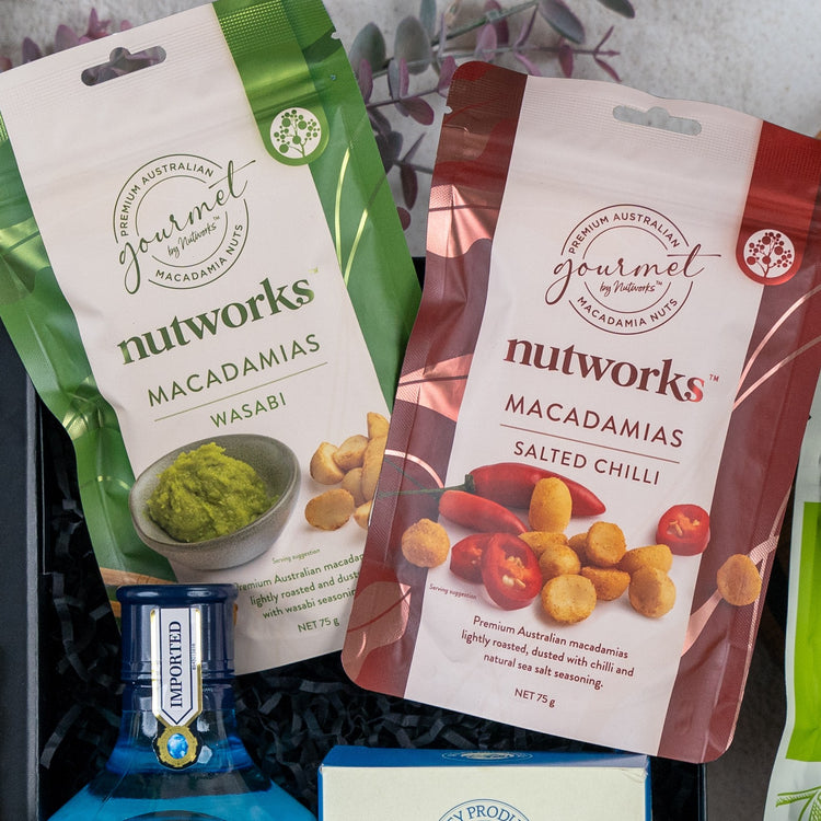 Two Nutworks macadamia nut packets—Wasabi (green) and Salted Chilli (red)—with images of seasoned nuts are displayed above a blue Bombay Sapphire bottle in The Hamper Boutique Co's Botanical Escape Hamper.