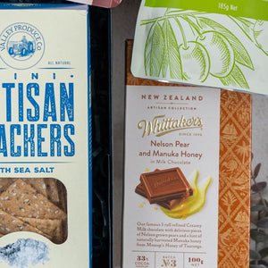 A close-up of three items from The Hamper Boutique Co's Bombay Sapphire Botanical Escape Hamper, including artisan crackers with sea salt, Whittakers Nelson Pear and Manuka Honey chocolate, and a partially visible green snack bag.