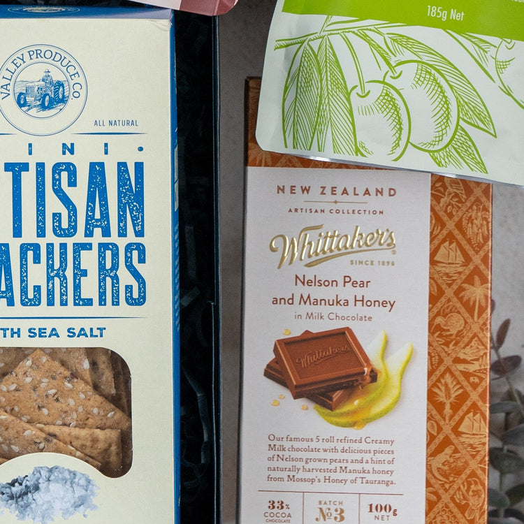 A close-up of three items from The Hamper Boutique Co's Bombay Sapphire Botanical Escape Hamper, including artisan crackers with sea salt, Whittakers Nelson Pear and Manuka Honey chocolate, and a partially visible green snack bag.