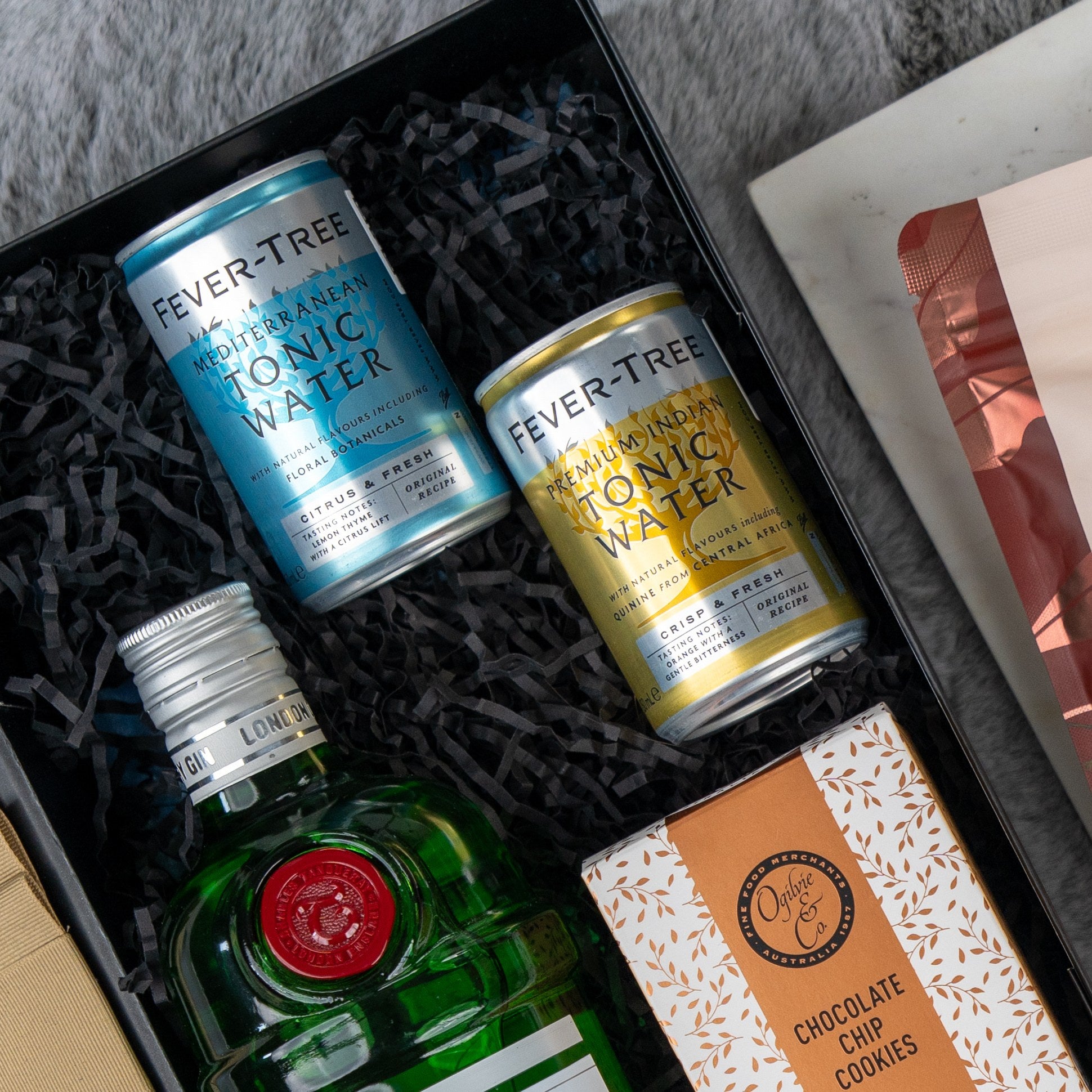 The Hamper Boutique Co’s Tanqueray Gin Journey Hamper includes Tanqueray gin, Fever-Tree tonic water cans, chocolate chip cookies, and other treats, all beautifully arranged on black crinkle paper—perfect for gifting.