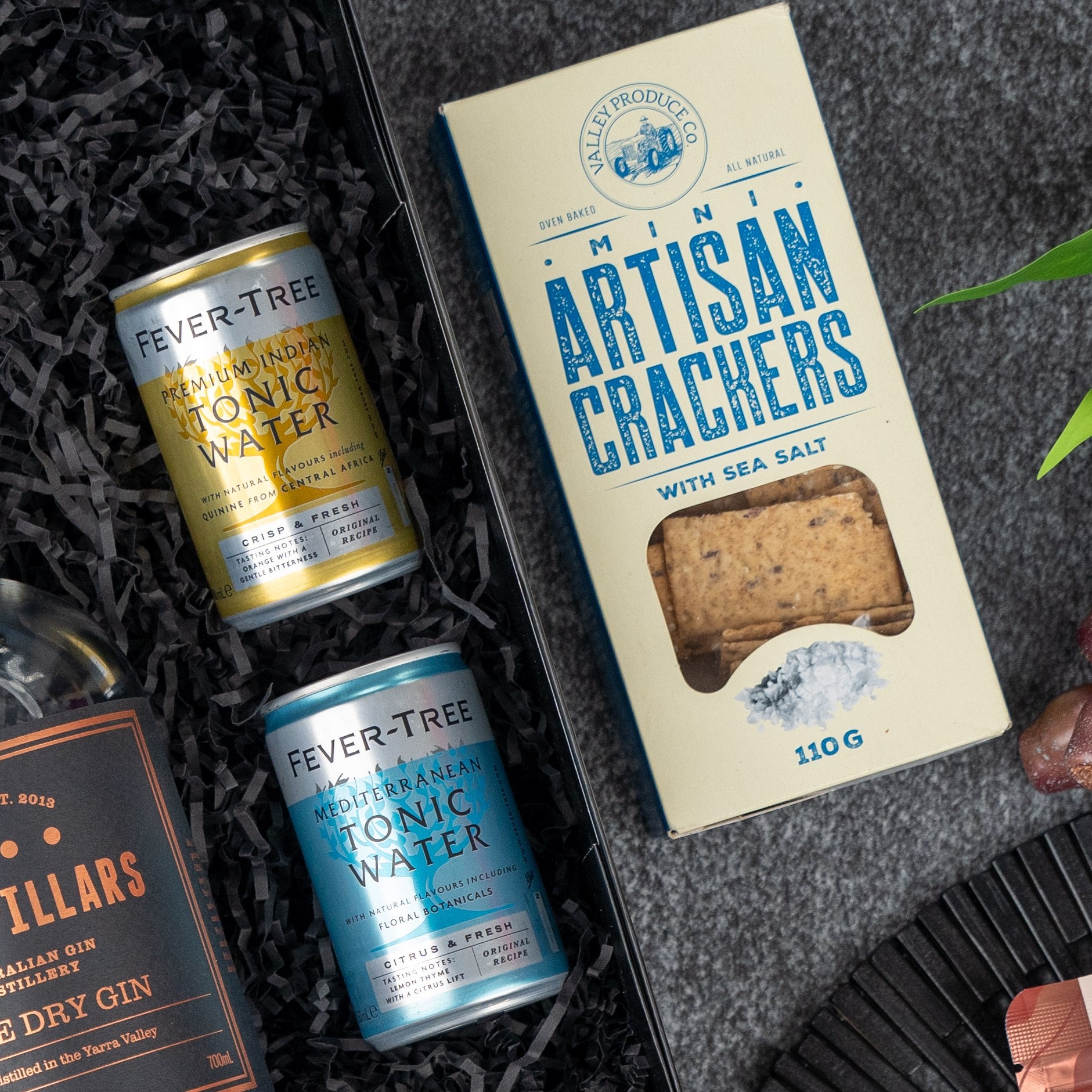 The Hamper Boutique Co’s Four Pillars Discovery Hamper includes Two Fever-Tree Mediterranean Tonic cans, a gin bottle, and sea salt Artisan Crackers, all beautifully presented on shredded black paper.