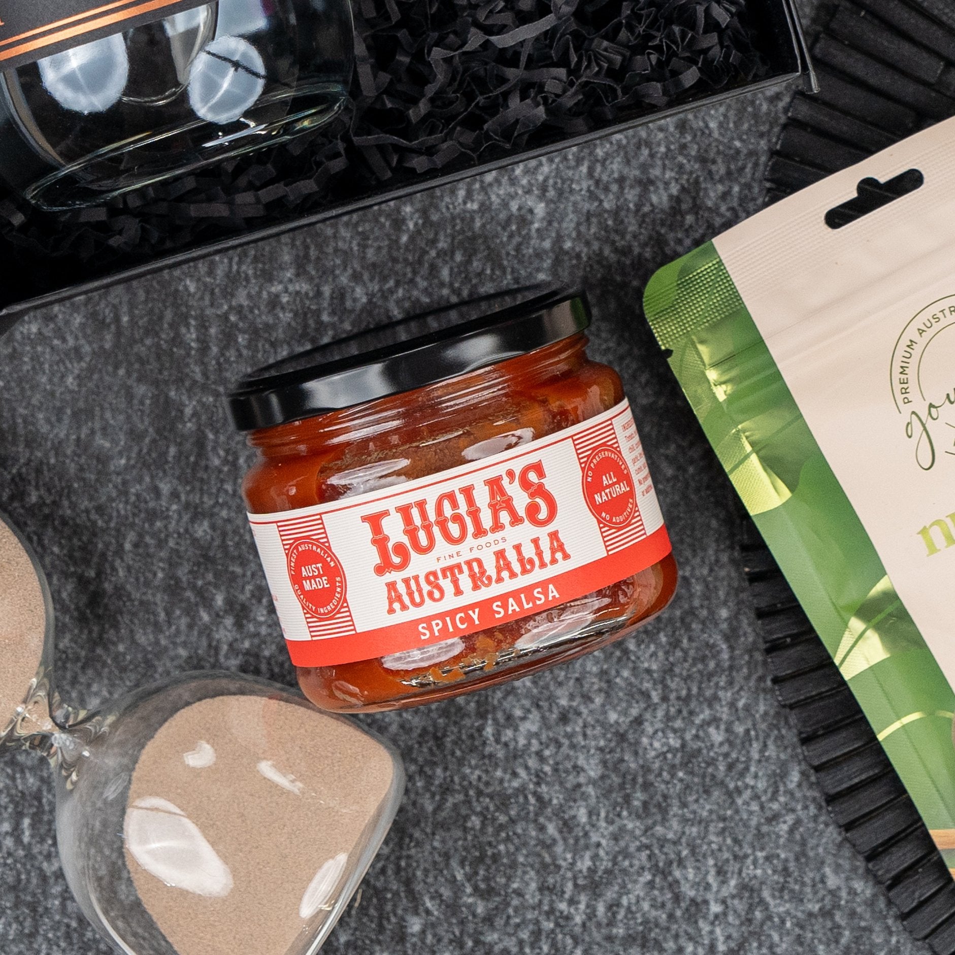 A Four Pillars Discovery Hamper by The Hamper Boutique Co sits on a gray surface, accompanied by a sand timer, a beige-green packet, and black shredded paper.