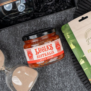 A Four Pillars Discovery Hamper by The Hamper Boutique Co sits on a gray surface, accompanied by a sand timer, a beige-green packet, and black shredded paper.