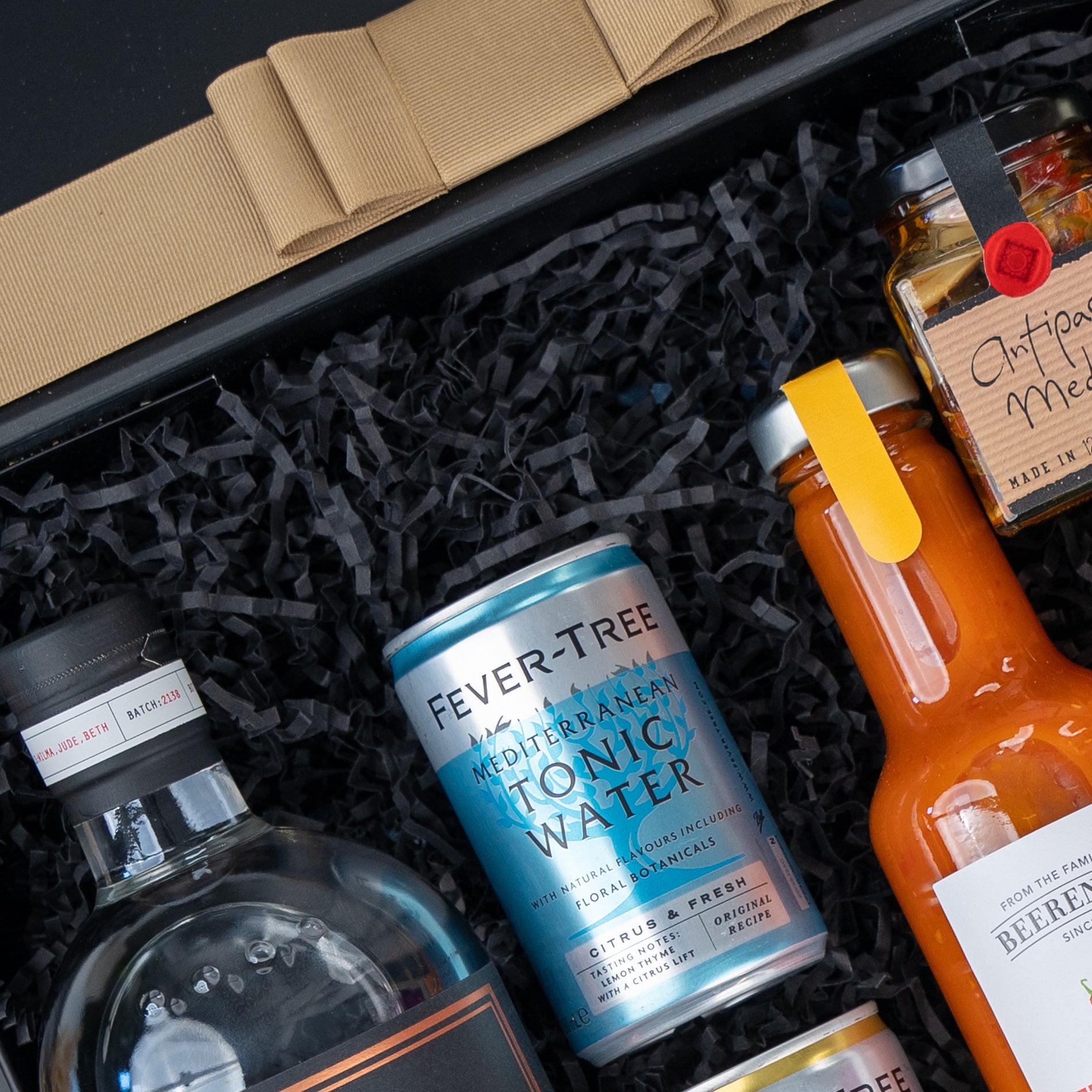 The Hamper Boutique Co Four Pillars Signature Collection Hamper includes a bottle of clear liquor, Fever-Tree Mediterranean Tonic Water, orange sauce, and Antipasto Misto in a gift box with black shredded paper.