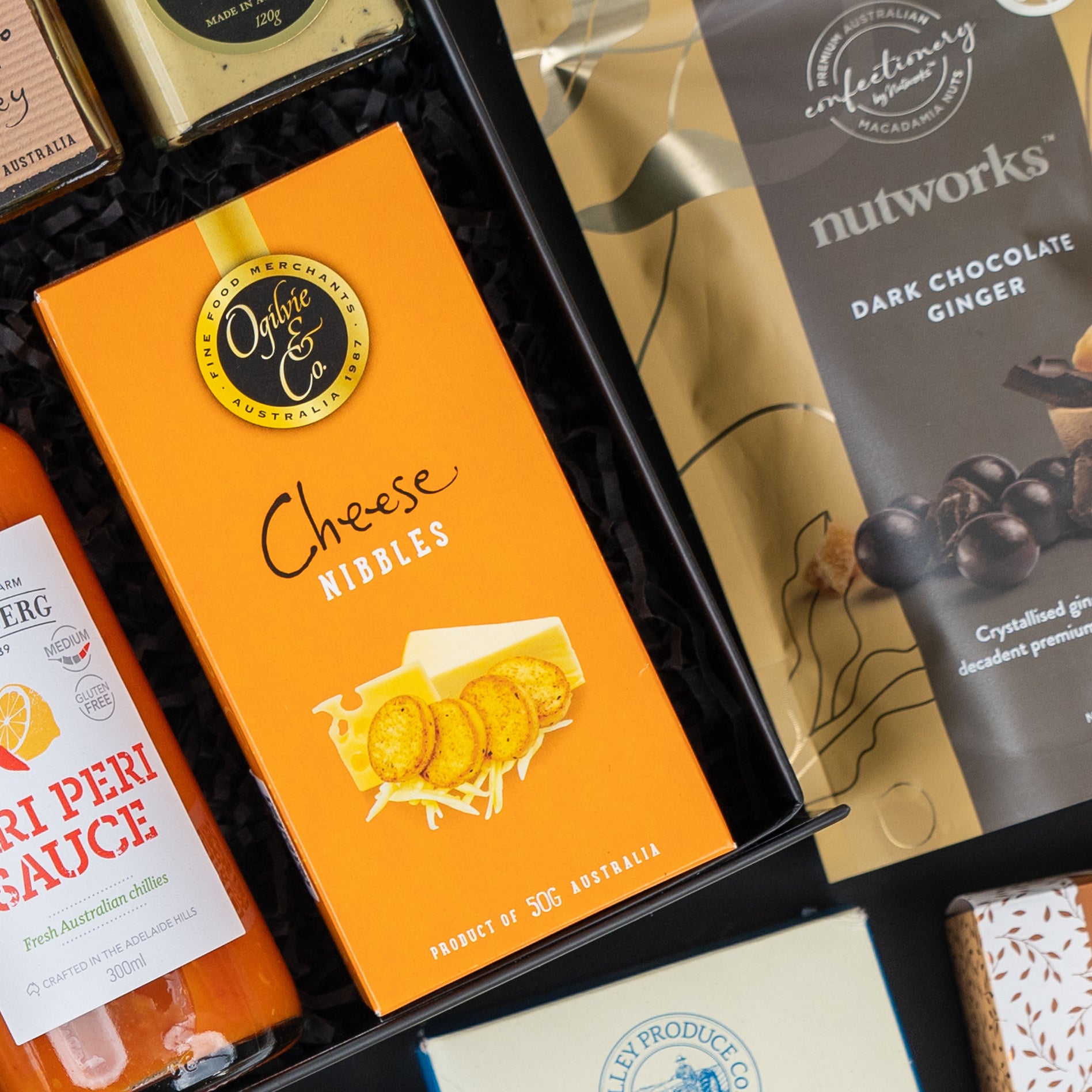 A close-up of The Hamper Boutique Co's Four Pillars Signature Collection Hamper, featuring a bright orange box of cheese nibbles, dark chocolate ginger, peri peri sauce, and assorted gourmet treats arranged neatly in a gift box.
