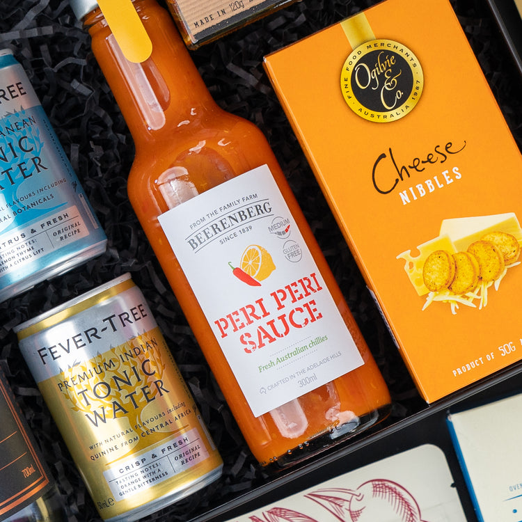 A close-up of The Hamper Boutique Co’s Four Pillars Signature Collection Hamper, featuring black shredded paper holding a bottle of sauce, Ogilvie & Co. Cheese Nibbles, and Fever-Tree Premium Indian Tonic Water.