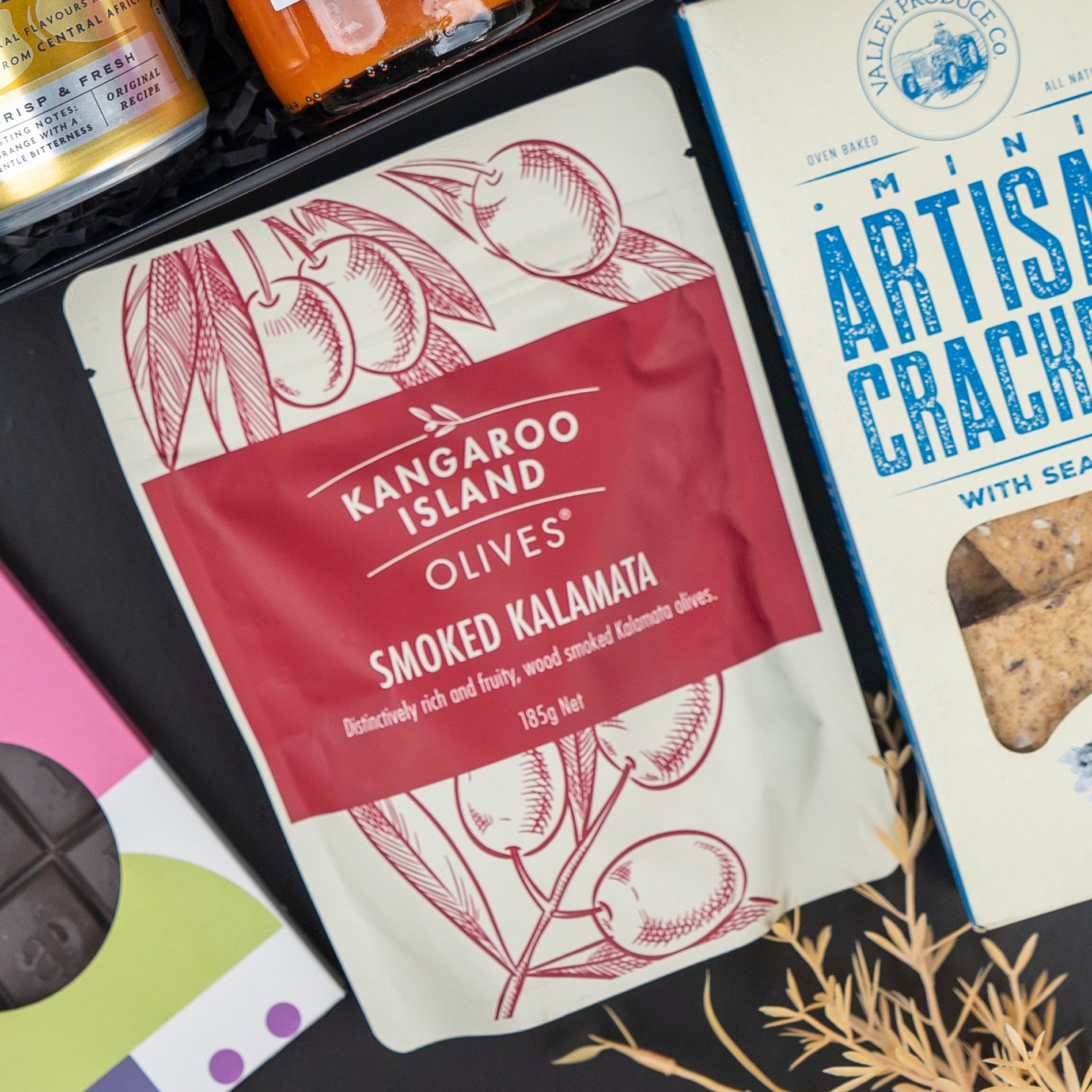 The Hamper Boutique Co’s Four Pillars Signature Collection Hamper features mini artisan sea salt crackers, Kangaroo Island Smoked Kalamata Olives, and other gourmet delights arranged on a dark surface.