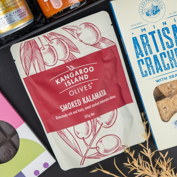The Hamper Boutique Co’s Four Pillars Signature Collection Hamper features mini artisan sea salt crackers, Kangaroo Island Smoked Kalamata Olives, and other gourmet delights arranged on a dark surface.