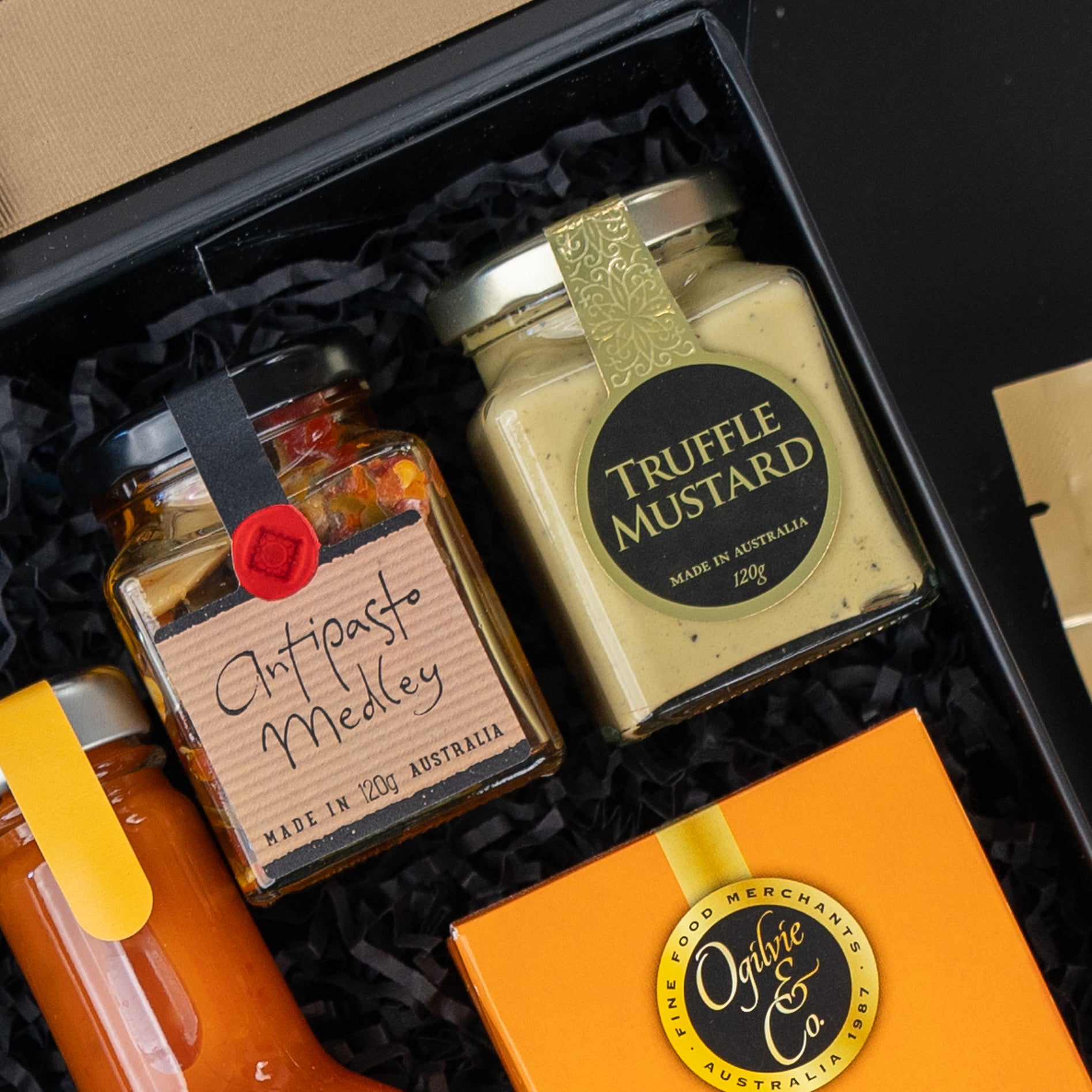 A close-up of The Hamper Boutique Co Four Pillars Signature Collection Hamper, featuring jars of antipasto medley and truffle mustard in an orange box, all arranged on black shredded paper.