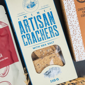 A box from The Hamper Boutique Co's Four Pillars Signature Collection Hamper, with a blue and white design and a window revealing mini artisan sea salt crackers, placed among other snack packages.