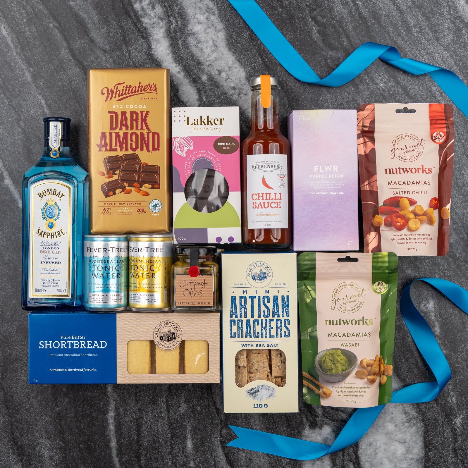A flat lay of The Hamper Boutique Co's Bombay Sapphire Indulgence Hamper, featuring Bombay Sapphire gin, chocolate, crackers, shortbread, nuts, tonic water, chili sauce in purple and blue boxes on dark marble with blue ribbons.