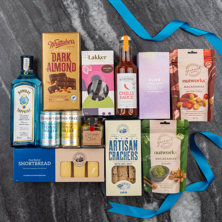 A flat lay of The Hamper Boutique Co's Bombay Sapphire Indulgence Hamper, featuring Bombay Sapphire gin, chocolate, crackers, shortbread, nuts, tonic water, chili sauce in purple and blue boxes on dark marble with blue ribbons.