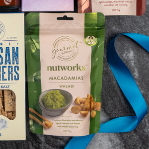 A Bombay Sapphire Indulgence Hamper from The Hamper Boutique Co is displayed on a gray countertop next to blue and white artisan crackers, a blue ribbon, and parts of other food packages.