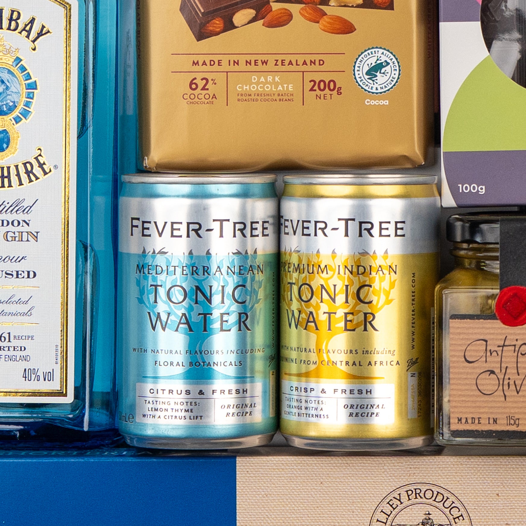 A close-up of The Hamper Boutique Co's Bombay Sapphire Indulgence Hamper, featuring gin, Fever-Tree tonic cans, dark chocolate, olives, and assorted gourmet treats beautifully arranged.