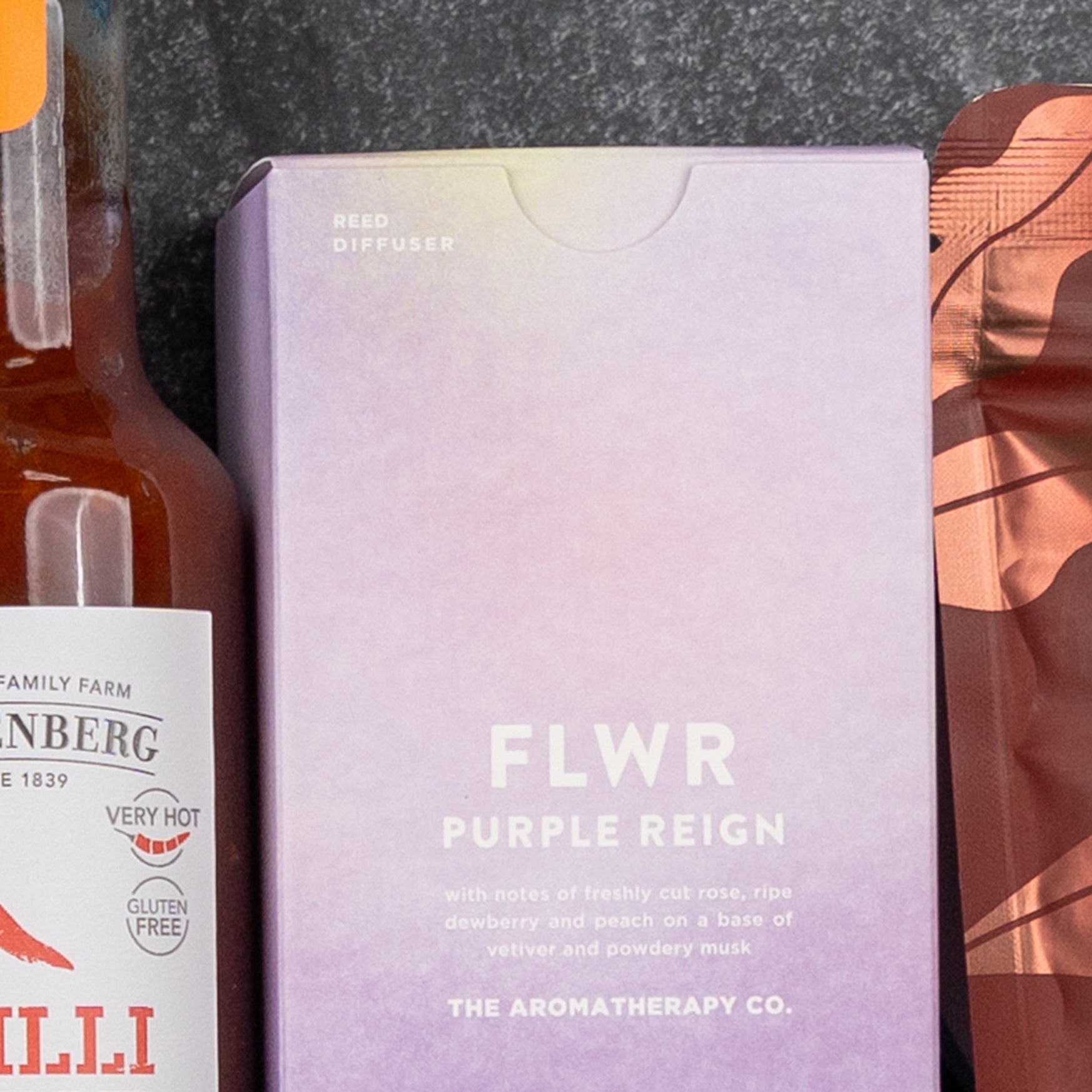 A close-up of three The Hamper Boutique Co Bombay Sapphire Indulgence Hamper packages: a glass bottle on the left, a lavender reed diffuser box labeled FLWR PURPLE REIGN in the center, and part of a metallic orange pouch on the right.