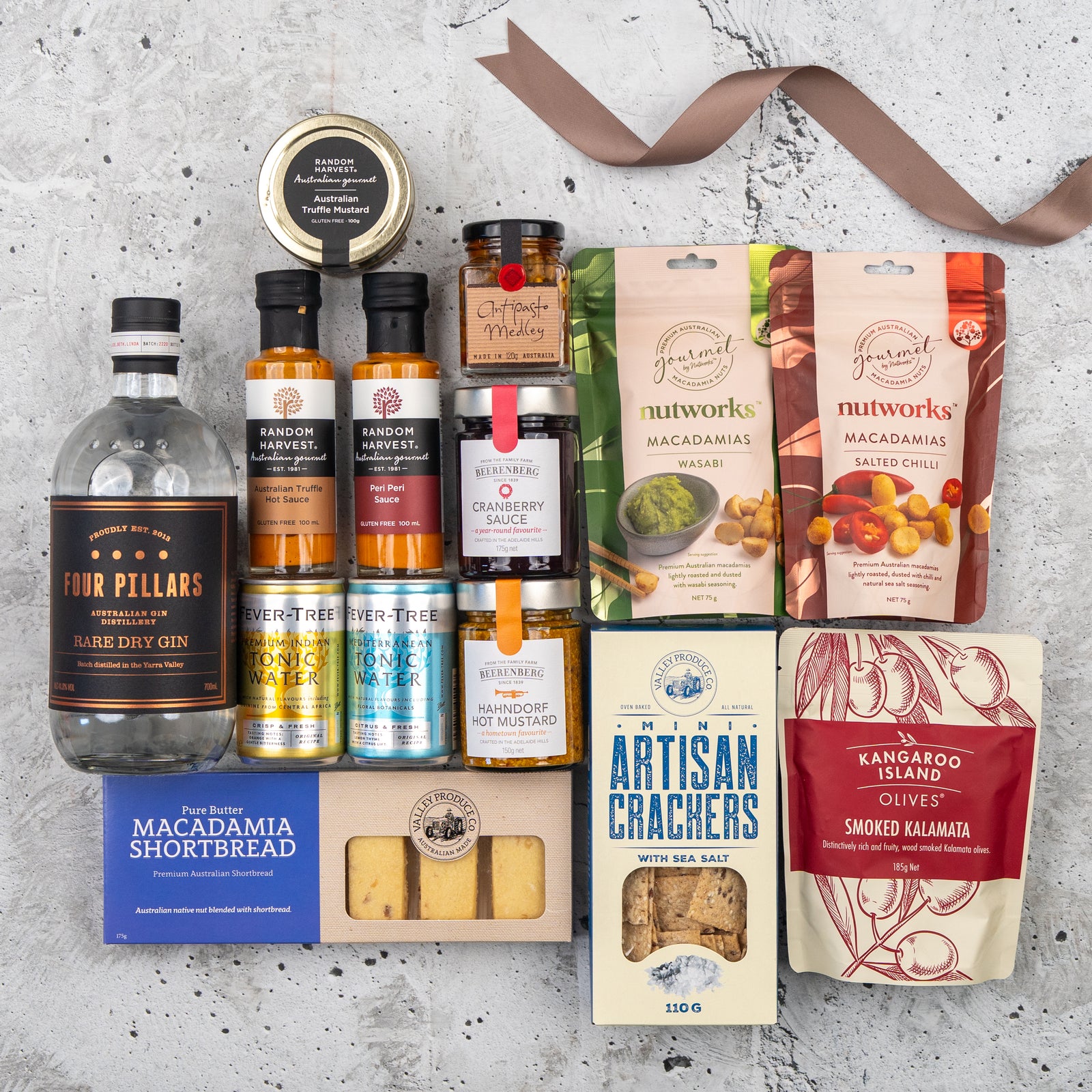 The Hamper Boutique Co's Four Pillars Premium Gin Hamper, arranged on a gray surface with a brown ribbon, features gin, ginger beer, chutneys, olives, macadamias, crackers, shortbread, sauces, and cranberry sauce.