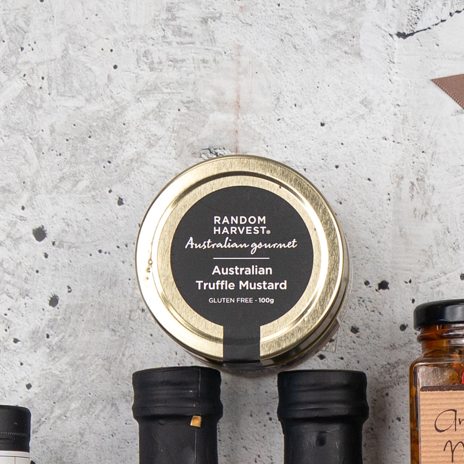 A top view of a jar labeled Four Pillars Premium Gin Hamper by The Hamper Boutique Co with a gold lid, placed on a textured gray surface next to bottles with black caps.