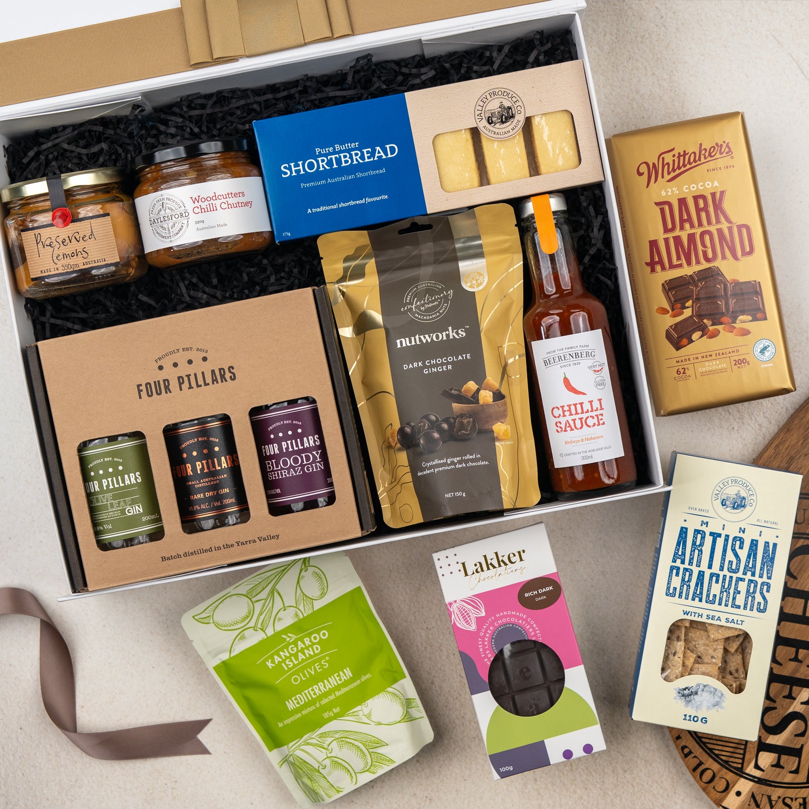 The Hamper Boutique Co’s Four Pillars Botanical Discovery Hamper features crackers, chocolate, shortbread, gin, jam, chutney, chilli sauce, and chocolate-coated nuts—beautifully presented on a light surface.