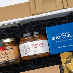 A close-up of The Hamper Boutique Co’s Four Pillars Botanical Discovery Hamper, featuring jars of preserved lemons, Woodcutters Chilli Chutney, and a blue box of pure butter shortbread nestled in black crinkle paper.