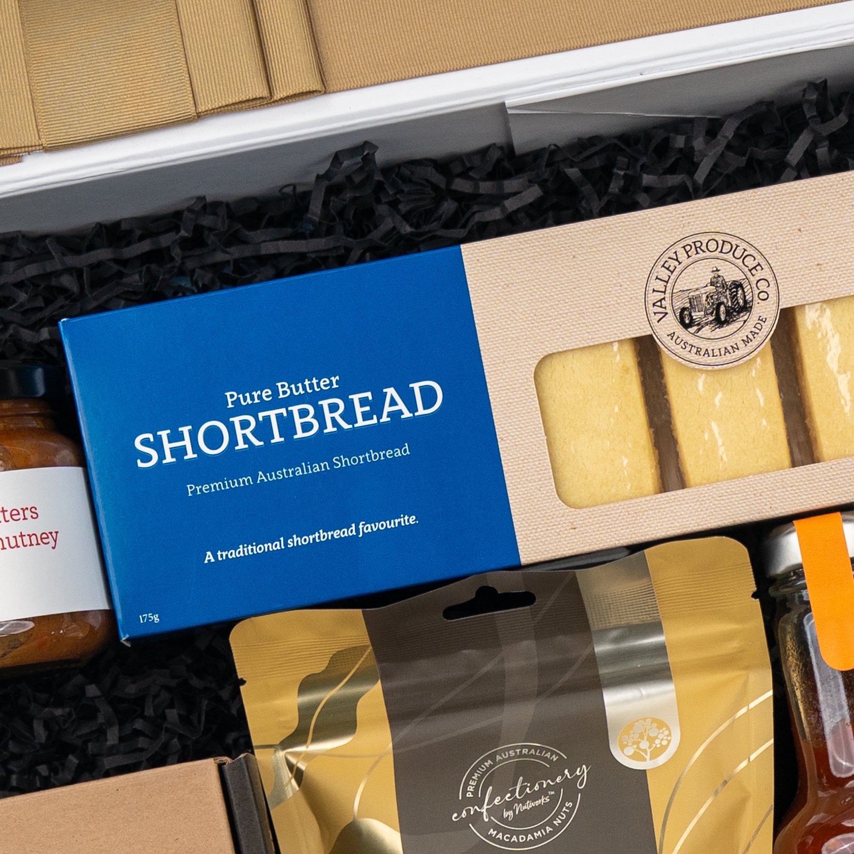 A box of Pure Butter Shortbread in blue packaging labeled Premium Australian Shortbread is included among the gourmet items in The Hamper Boutique Co's Four Pillars Botanical Discovery Hamper, nestled in black shredded paper.