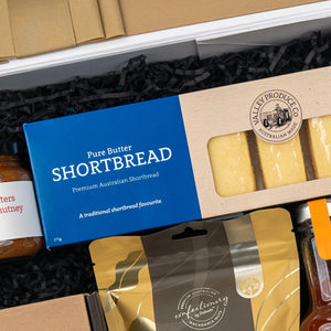 A box of Pure Butter Shortbread in blue packaging labeled Premium Australian Shortbread is included among the gourmet items in The Hamper Boutique Co's Four Pillars Botanical Discovery Hamper, nestled in black shredded paper.