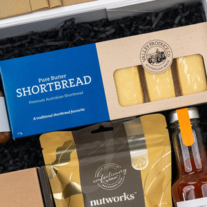 The Hamper Boutique Co’s Four Pillars Botanical Discovery Hamper includes pure butter shortbread, macadamia nut mix, and a bottle of sauce in a blue box, all elegantly arranged on black shredded paper.