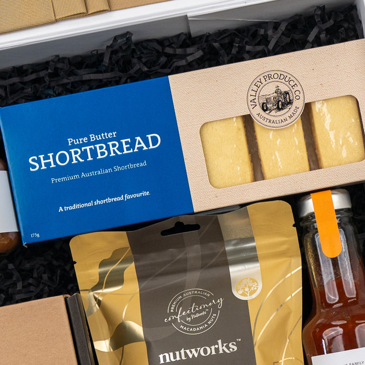 The Hamper Boutique Co’s Four Pillars Botanical Discovery Hamper includes pure butter shortbread, macadamia nut mix, and a bottle of sauce in a blue box, all elegantly arranged on black shredded paper.