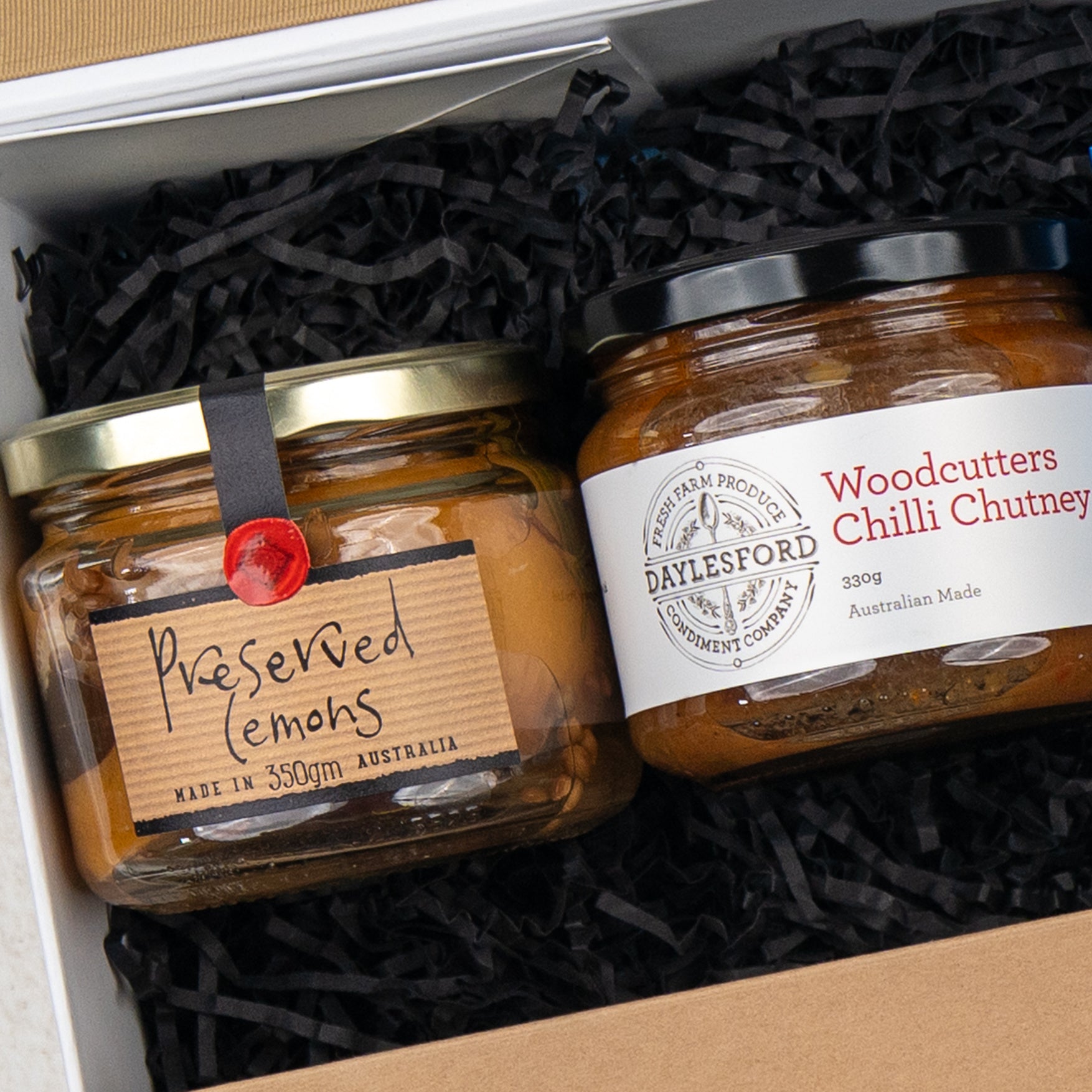 The Four Pillars Botanical Discovery Hamper by The Hamper Boutique Co includes two glass jars: Preserved Lemons with a handwritten tag and Woodcutters Chilli Chutney with a printed label, packed in shredded black paper. Both products are from Australia.